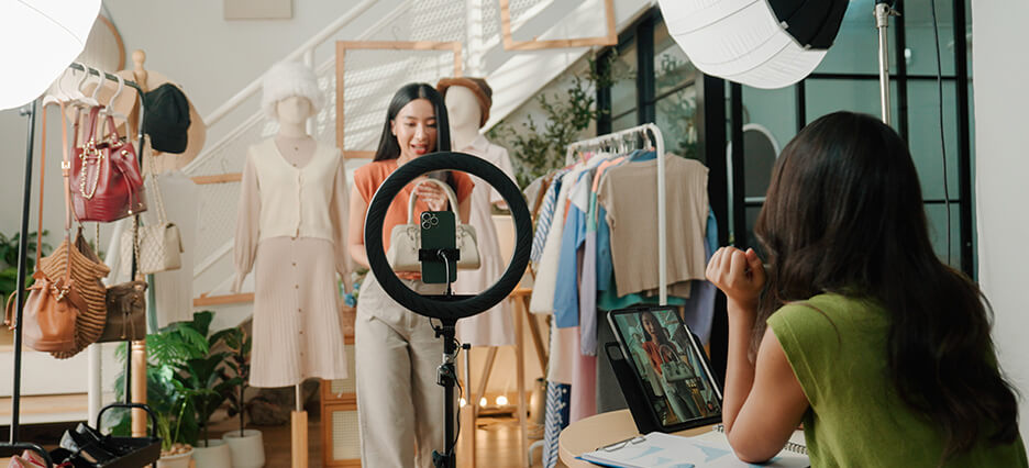 A young Asian woman live-selling fashion items to online customers while her friend is managing the broadcast behind the scene